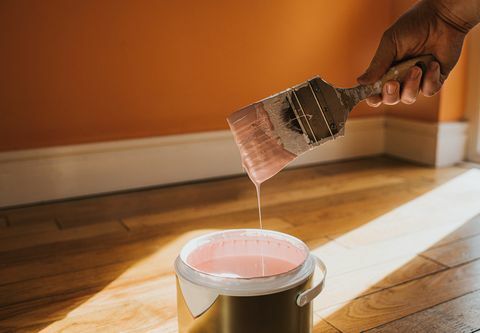 la mano sostiene un pincel grueso y lo quita, después de sumergirlo en la pintura crea una gota de pintura que salpica De vuelta a la olla, la pared de atrás está pintada de naranja y el color de la lata es rosa, lo que choca conceptual con el espacio para Copiar