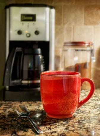 Taza de café humeante: Una taza roja de café humeante descansa sobre una encimera de cocina de granito. En el fondo se ve una cafetera y un recipiente con café molido.